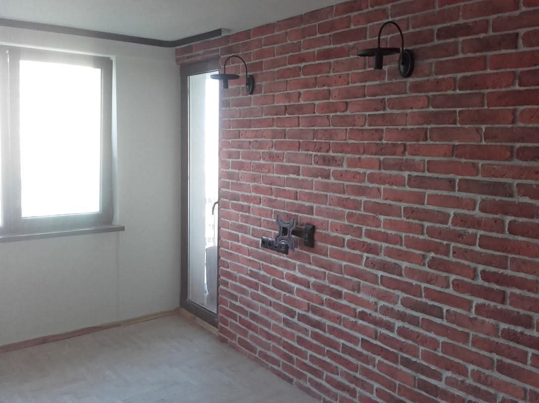 Balkon po remoncie: ściana z cegły, oświetlenie, uchwyt TV. Widok na okno i drzwi balkonowe. Jasna podłoga. Styl industrialny, nowoczesny.