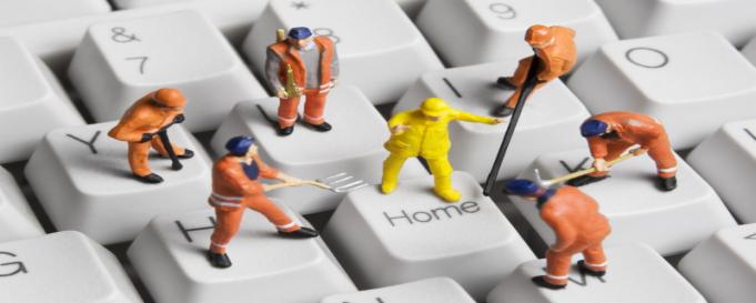 Miniaturowe figurki robotników w kombinezonach naprawiające klawiaturę komputera, jeden stoi na klawiszu 'Home' w żółtym kombinezonie.
