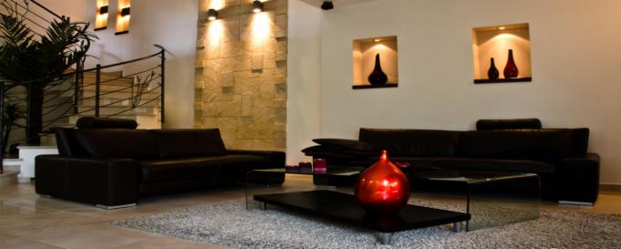 Nowoczesny salon z czarnymi skórzanymi sofami, szklanymi stolikami kawowymi i ścianą z kamienia dekoracyjnego podświetloną punktowymi lampami. Czerwona ozdobna kula na stoliku kawowym. W tle schody...