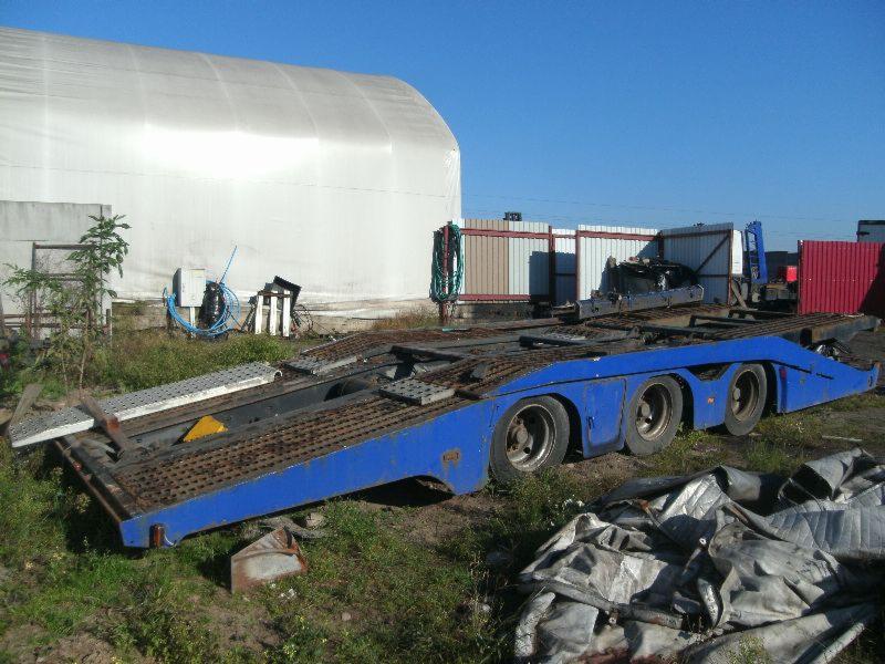 Niebieska, trzyosiowa laweta z najazdami na tle magazynu z plandeki i ogrodzenia, widoczny sprzęt i plandeki na ziemi.