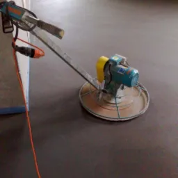 Wyrównywanie wylewki samopoziomującej za pomocą elektrycznej zacieraczki do betonu. Widoczne okno w tle i pomarańczowy kabel zasilający.