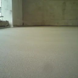 Wyrównana, szara wylewka podłogowa w surowym wnętrzu z fragmentem okna i widoczną ścianą. Gładka powierzchnia przygotowana do dalszych prac wykończeniowych.