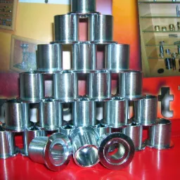 Metalowe tuleje ułożone w piramidę na czerwonym tle, obok szyldu firmy z branży obróbki metali. Błyszczące powierzchnie, precyzyjne wykonanie.