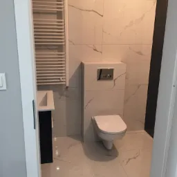 Nowoczesna łazienka z marmurowymi płytkami, wiszącą toaletą, grzejnikiem drabinkowym i czarną szafką pod umywalką.