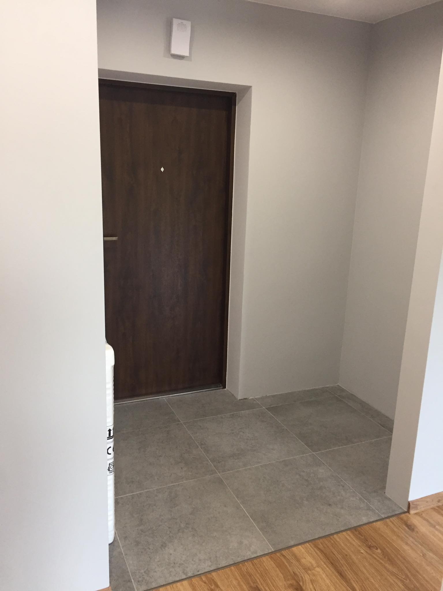 Wnętrze przedpokoju z ciemnymi drzwiami wejściowymi, szarą podłogą z płytek i jasnymi ścianami, widoczny fragment podłogi z paneli oraz opakowanie materiałów budowlanych.