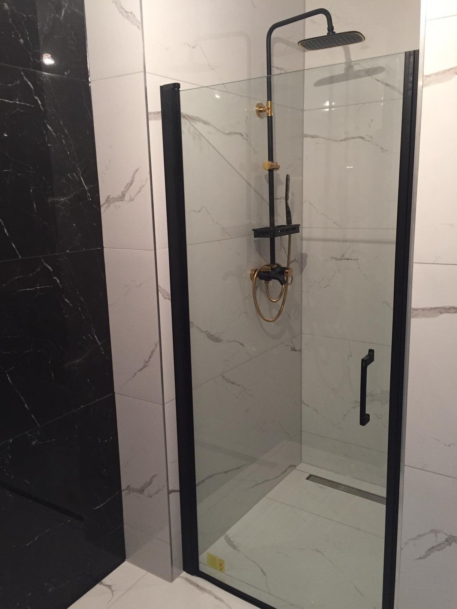Kabina prysznicowa z czarnymi ramami i złotymi elementami armatury na tle białych płytek imitujących marmur, z fragmentem ściany wyłożonej czarnym marmurem.