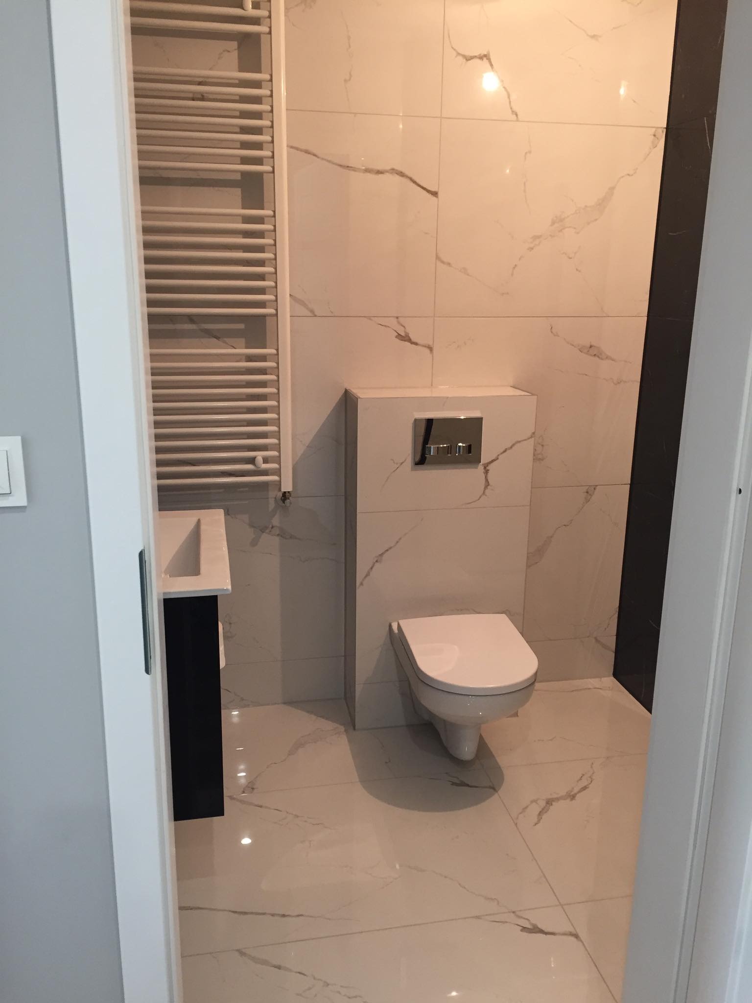 Nowoczesna łazienka z marmurowymi płytkami, wiszącą toaletą, grzejnikiem drabinkowym i czarną szafką pod umywalką.