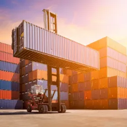 Czerwony wózek widłowy typu reach stacker podnosi kontener na tle stosu kontenerów w porcie, oświetlony ciepłym światłem zachodzącego słońca.