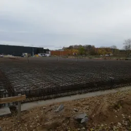 Rozległy plac budowy z przygotowanym zbrojeniem fundamentów pod budynek, widoczne szalunki i betonowe wylewki obwodowe, w tle budynek magazynowy i drzewa.