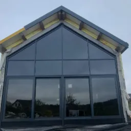 Nowoczesna fasada budynku z dużymi, grafitowymi oknami aluminiowymi, widok z zewnątrz, w tle zabudowa jednorodzinna, pochmurne niebo.