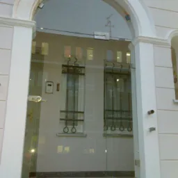 Szklane drzwi wejściowe z łukowym nadprożem, widoczne elementy mocujące szkło i odblask wnętrza budynku z oknami z ozdobnymi kratami.