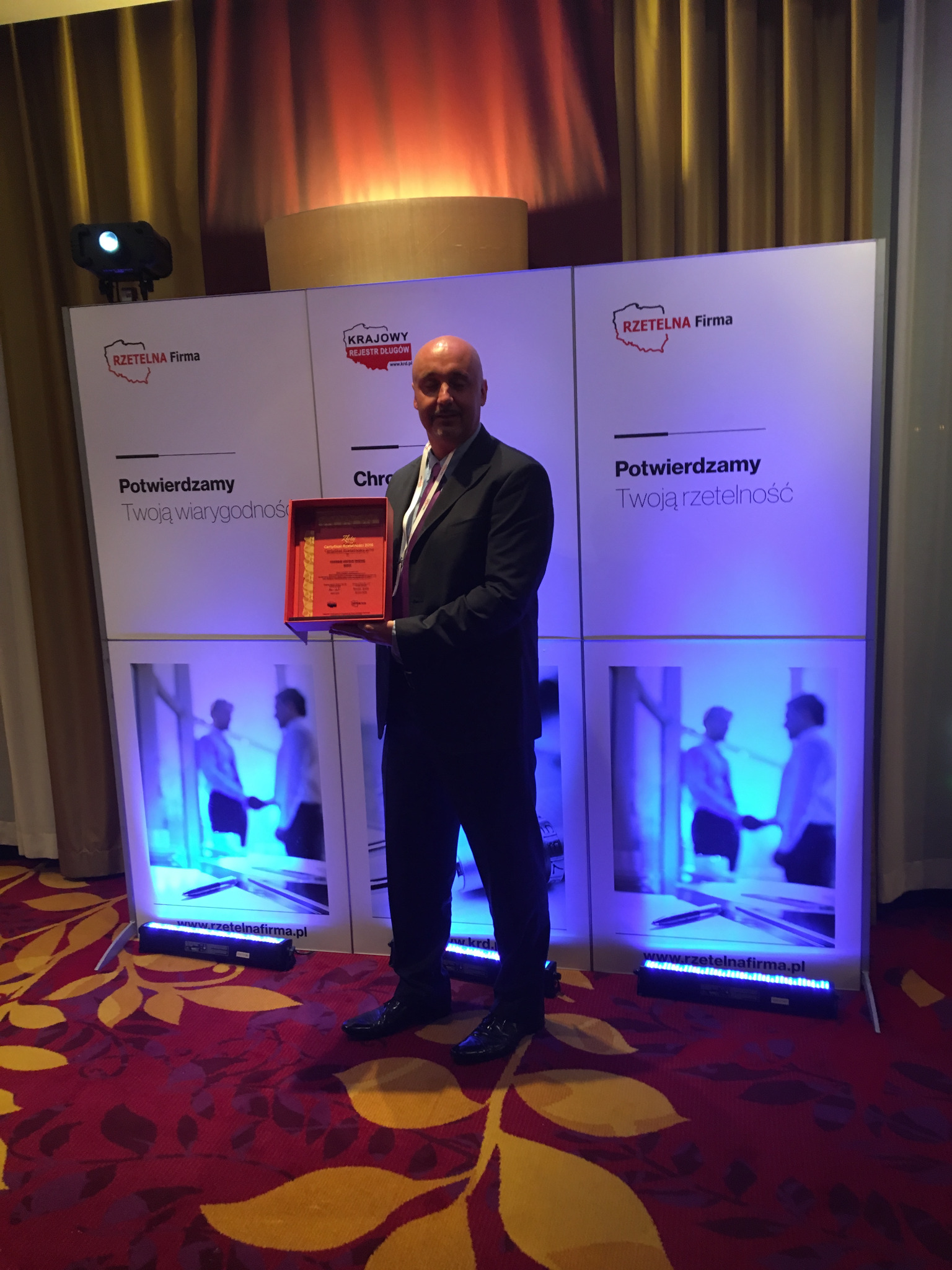 Mężczyzna w garniturze prezentuje certyfikat 'Rzetelna Firma' na tle ścianki reklamowej z logo i hasłem 'Potwierdzamy Twoją wiarygodność/rzetelność', podświetlonej niebieskim światłem.