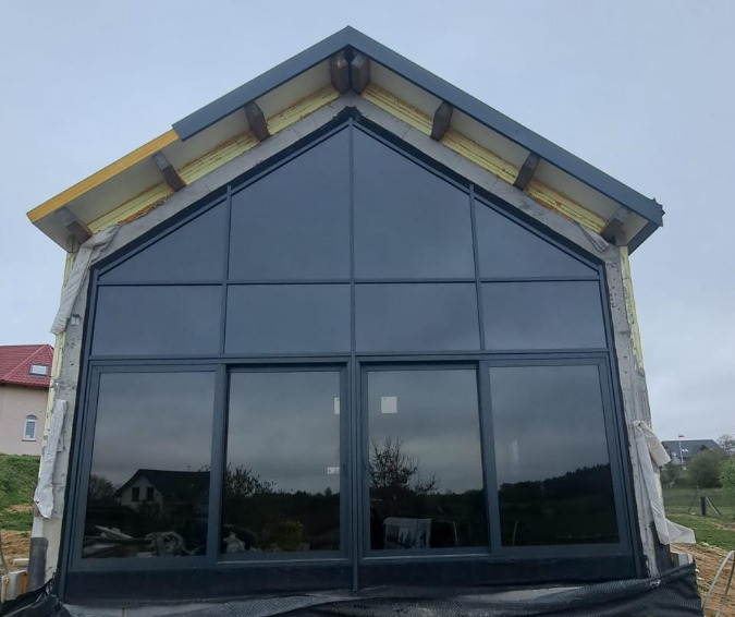 Nowoczesna fasada budynku z dużymi, grafitowymi oknami aluminiowymi, widok z zewnątrz, w tle zabudowa jednorodzinna, pochmurne niebo.