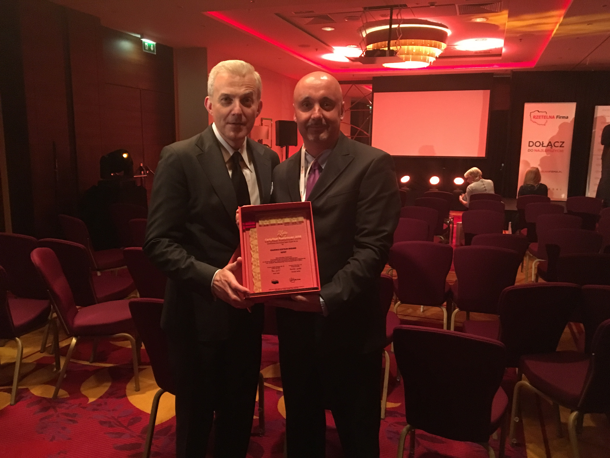Dwóch mężczyzn w garniturach prezentuje certyfikat w sali konferencyjnej z czerwonymi krzesłami i oświetleniem. W tle widoczny ekran i banery.