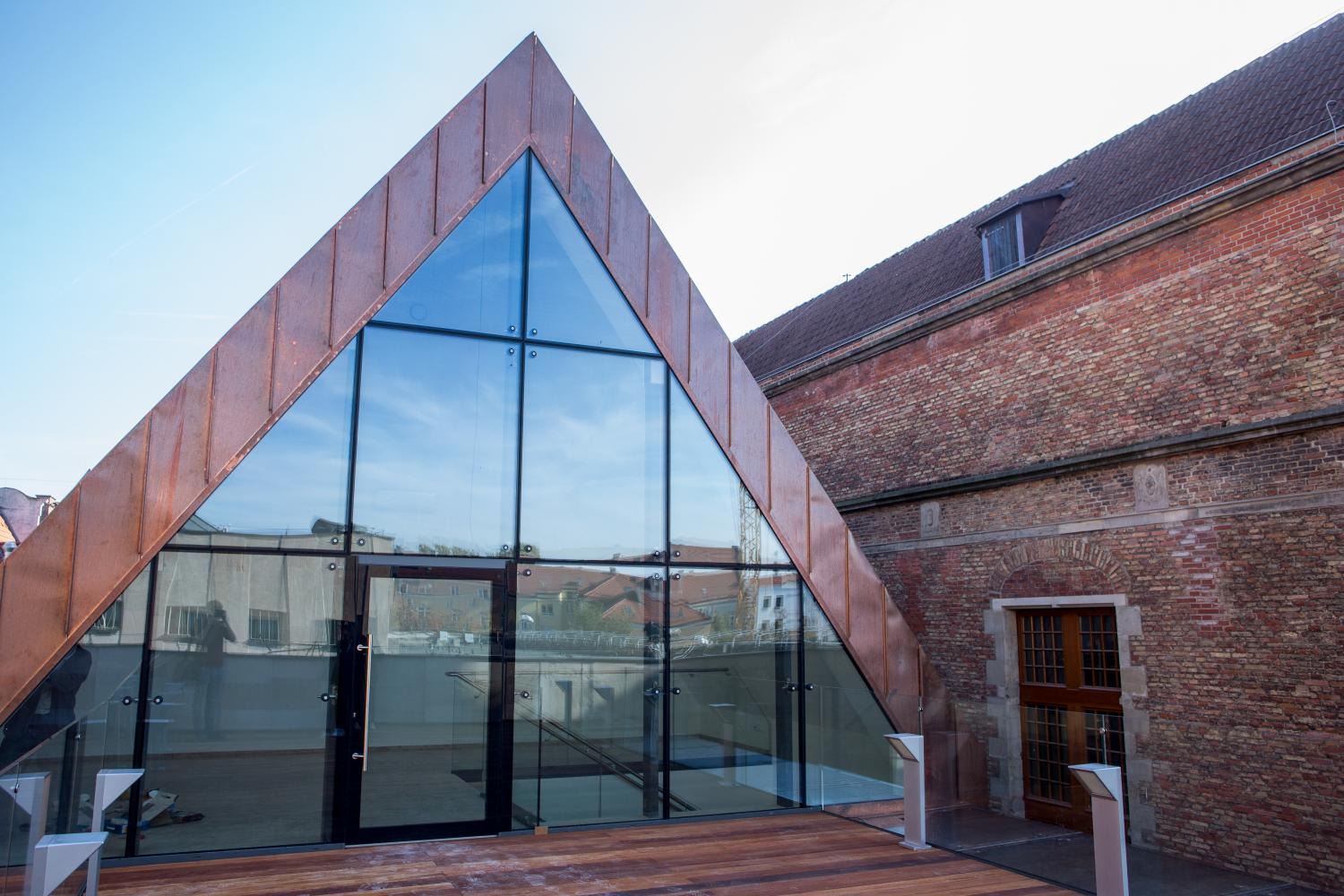 Nowoczesna szklana fasada budynku w kształcie trójkąta z metalowym obramowaniem w kolorze miedzi, przylegająca do ceglanego budynku z drewnianymi drzwiami.