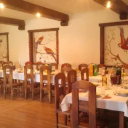 Uroczysta sala z długim stołem nakrytym białym obrusem, drewnianymi krzesłami i dekoracjami. Na ścianach wiszą obrazy z motywem papug. Widoczne drewniane belki na suficie.