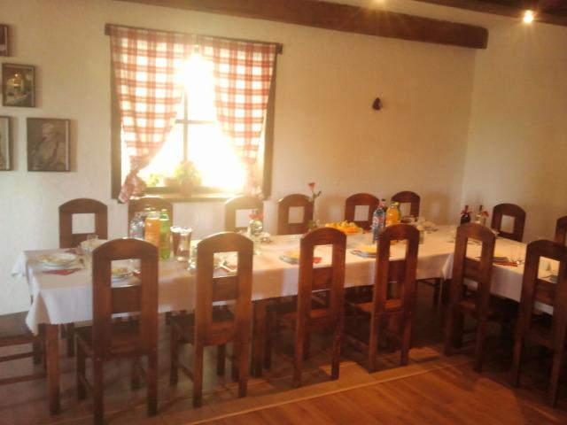 Sala jadalna w rustykalnym stylu z długim stołem nakrytym białym obrusem, drewnianymi krzesłami i napojami. W tle okno z czerwoną kratą i dekoracje.