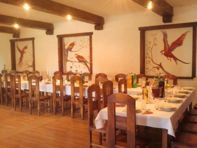 Uroczysta sala z długim stołem nakrytym białym obrusem, drewnianymi krzesłami i dekoracjami. Na ścianach wiszą obrazy z motywem papug. Widoczne drewniane belki na suficie.