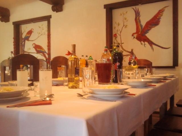 Ustawiony stół z białym obrusem, nakrycia, napoje i dekoracje. W tle obrazy z motywem papug w drewnianych ramach. Przygotowania do uroczystości.