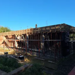 Wznoszenie ścian parterowego budynku z czerwonej cegły, widoczne szalunki i podpory, teren budowy częściowo zalany wodą, na górze konstrukcji stoi człowiek.