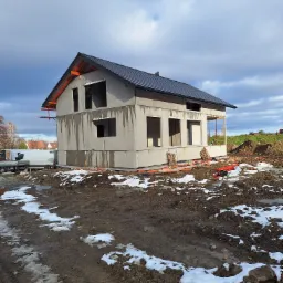 Surowy stan domu murowanego z czarnym dachem, widoczne otwory okienne bez okien, częściowo ocieplone ściany zewnętrzne, teren budowy z błotem i resztkami śniegu, w tle zaparkowany biały bus.