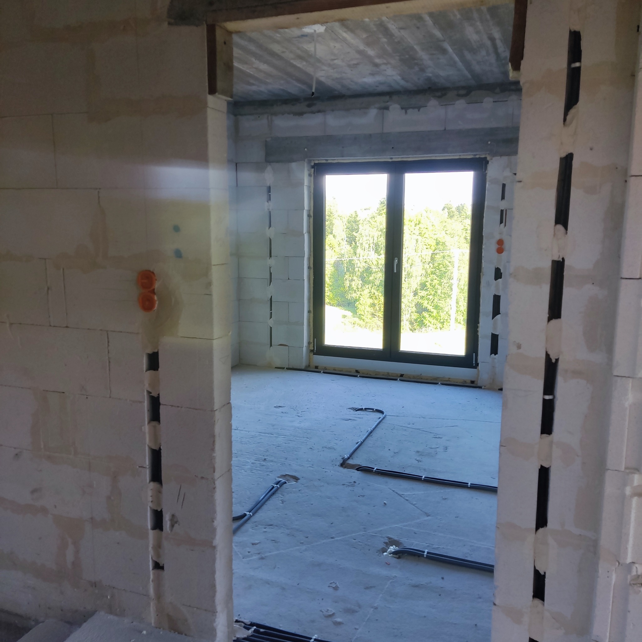 Wnętrze w surowym stanie z widocznymi kablami elektrycznymi ułożonymi na podłodze i w ścianach. Pomarańczowe puszki pod gniazdka osadzone w ścianie z betonu komórkowego.