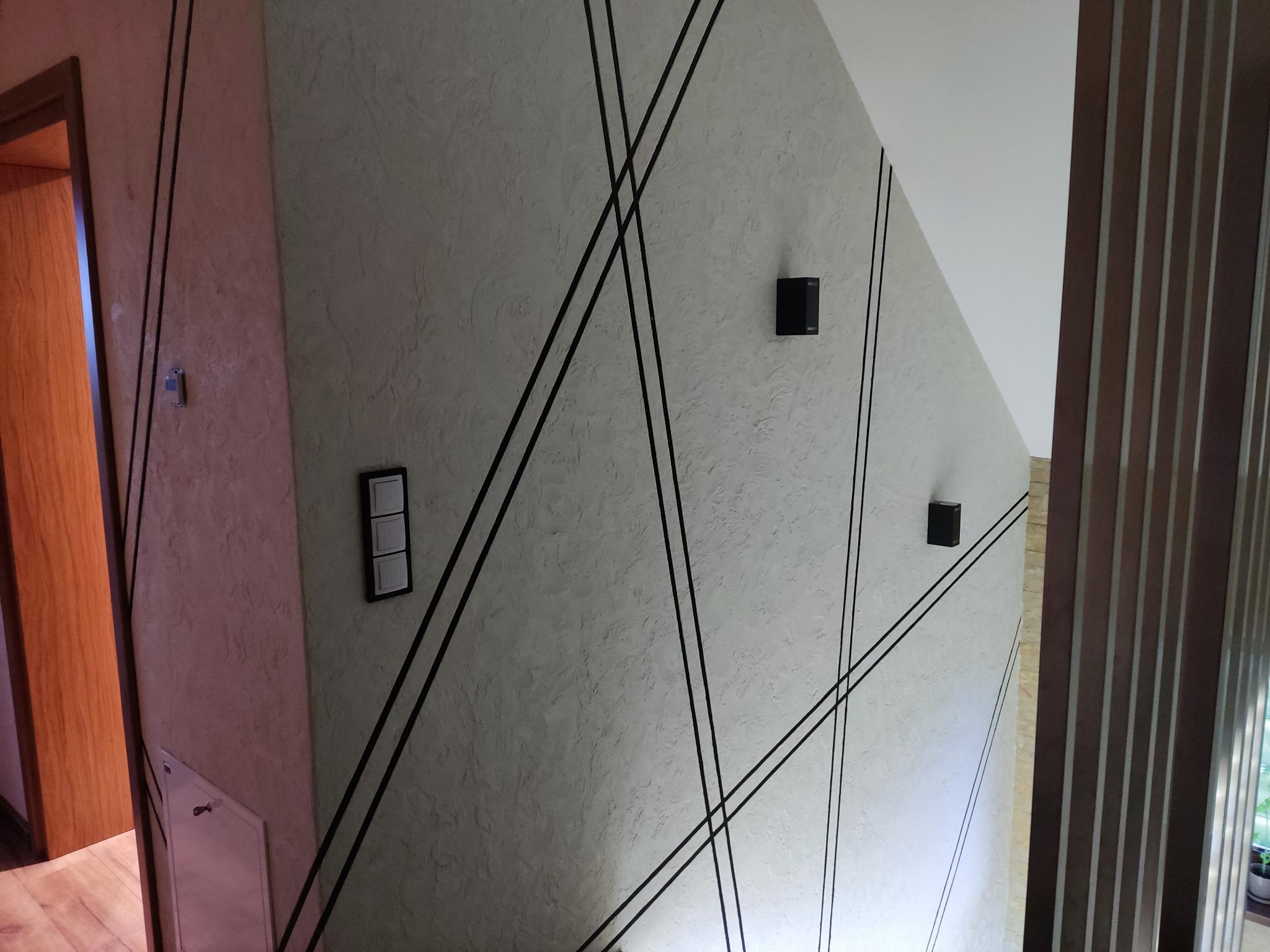 Ściana z dekoracyjnym tynkiem, czarnymi listwami, włącznikiem i czujnikami. Elementy instalacji elektrycznej wkomponowane w design wnętrza.