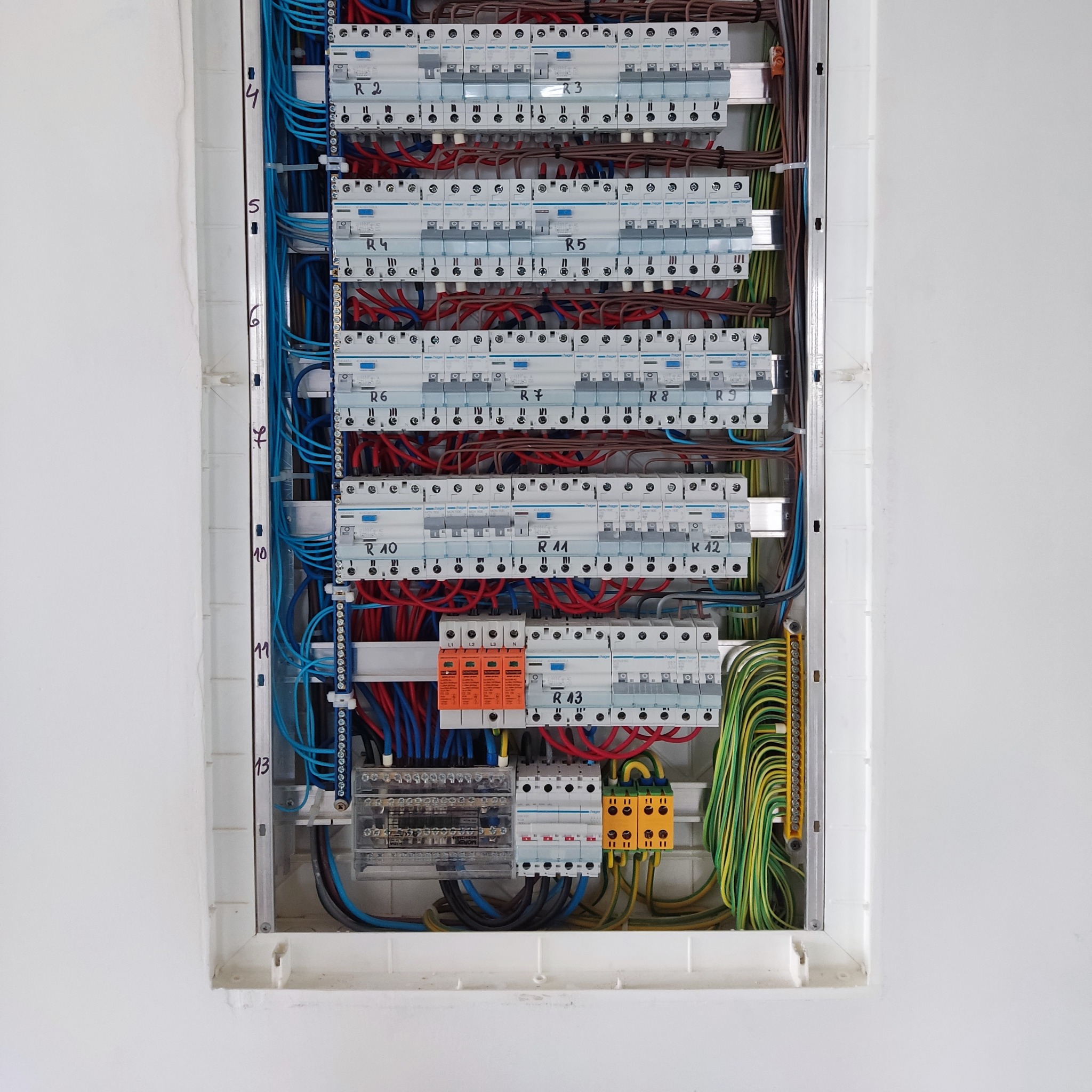 Rozdzielnia elektryczna w białej obudowie z numeracją przewodów na ścianie, widoczne bezpieczniki i okablowanie w kolorach: niebieskim, czerwonym, zielono-żółtym.
