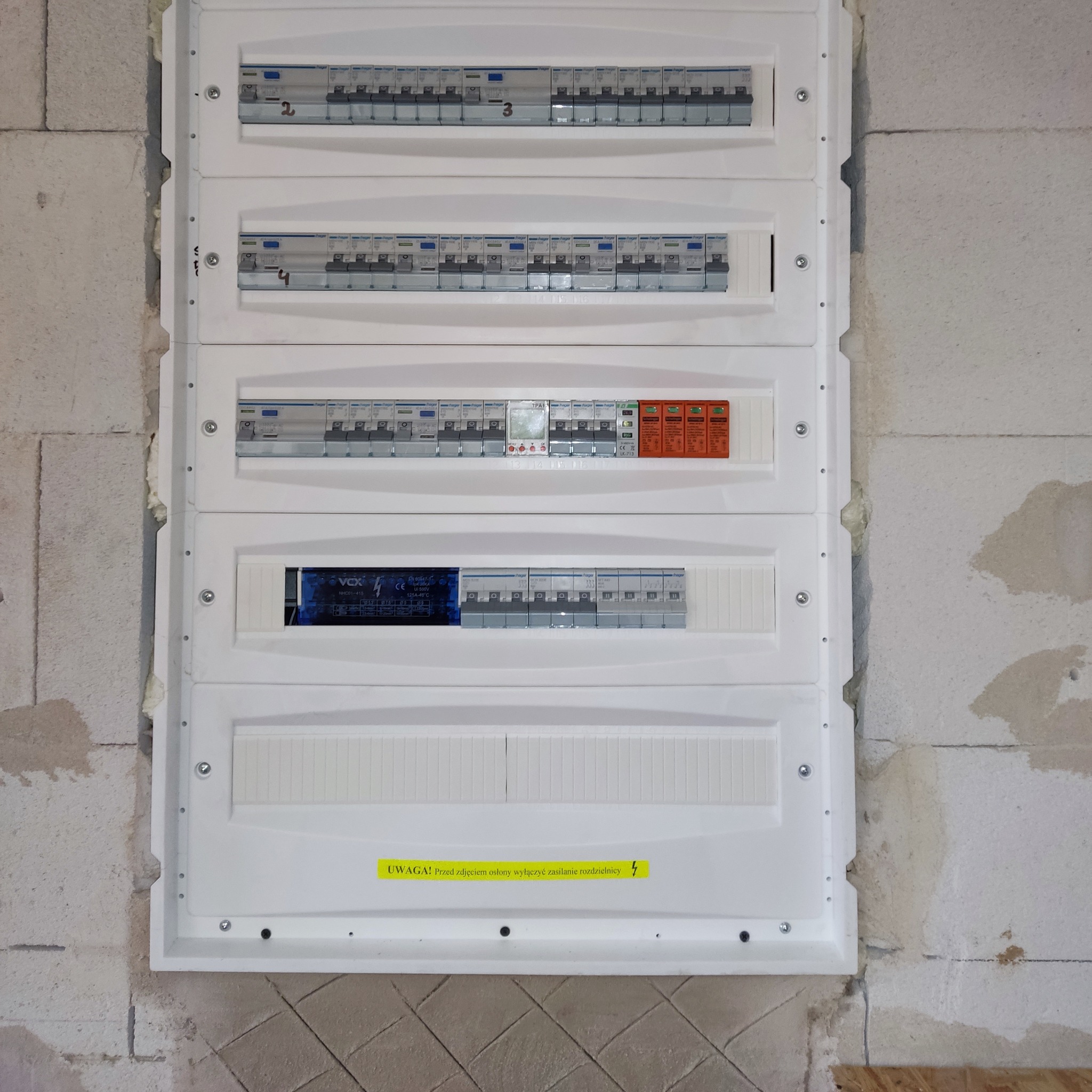 Rozdzielnica elektryczna natynkowa, zamontowana w surowym pomieszczeniu z widoczną strukturą bloczków. Widoczne zabezpieczenia, bezpieczniki i ostrzegawcza naklejka.