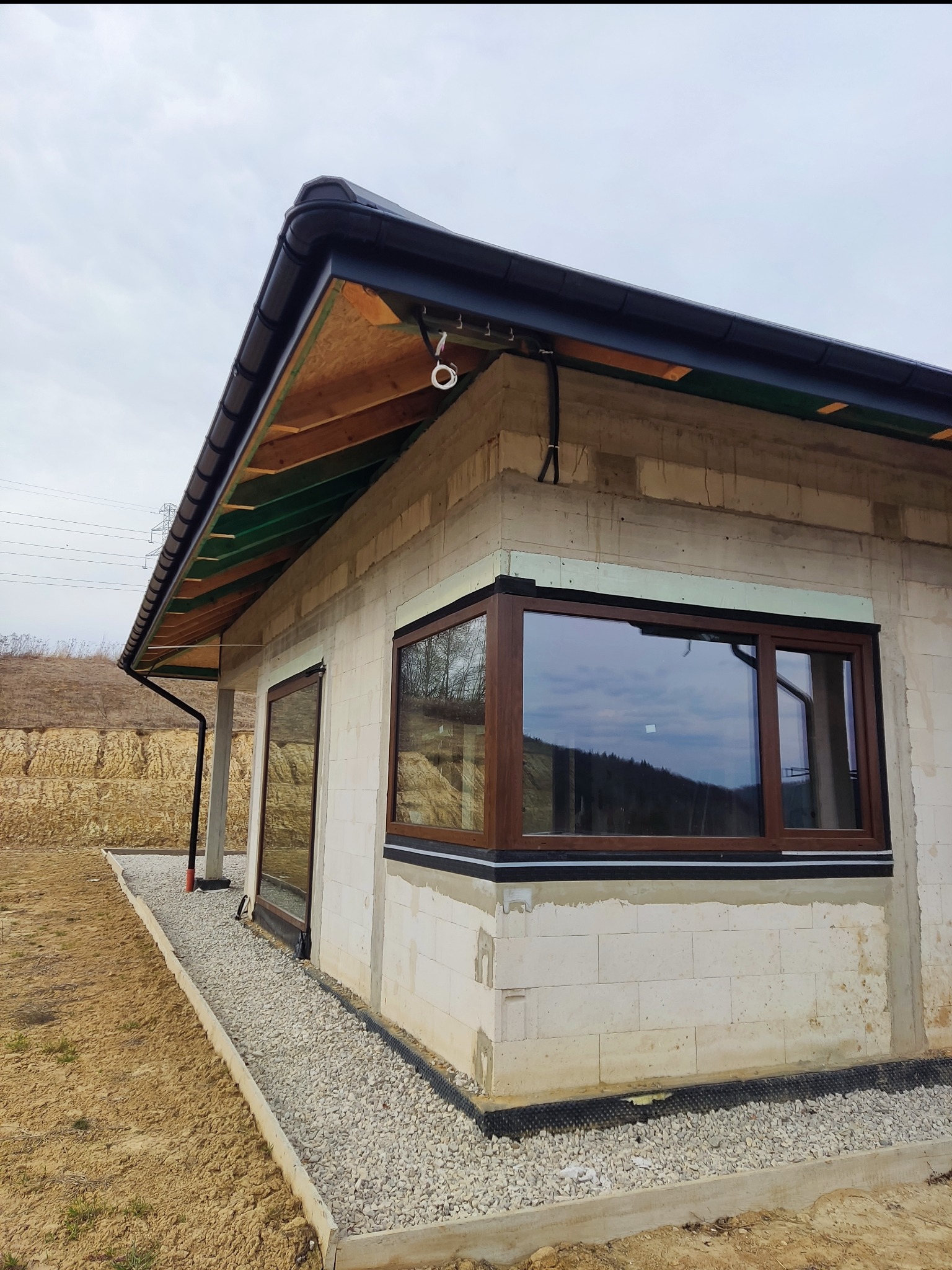 Niedokończony budynek z widocznymi kablami elektrycznymi na elewacji. Okna z brązowymi ramami, dach z czarną blachą. W tle ziemny nasyp.