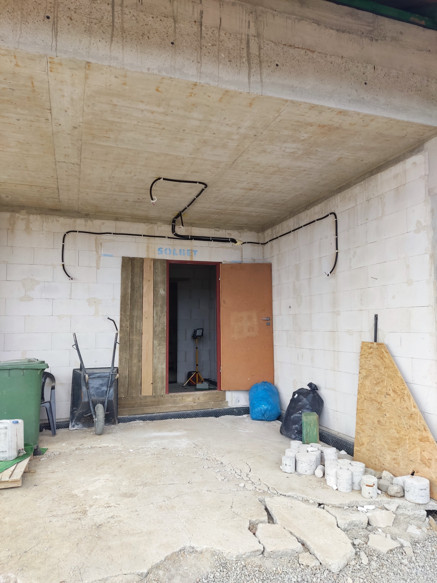 Wnętrze w surowym stanie z widocznymi przewodami podtynkowymi. Na ścianie napis SOLBET. W tle widać lampę budowlaną. Widoczny gruz i materiały budowlane.