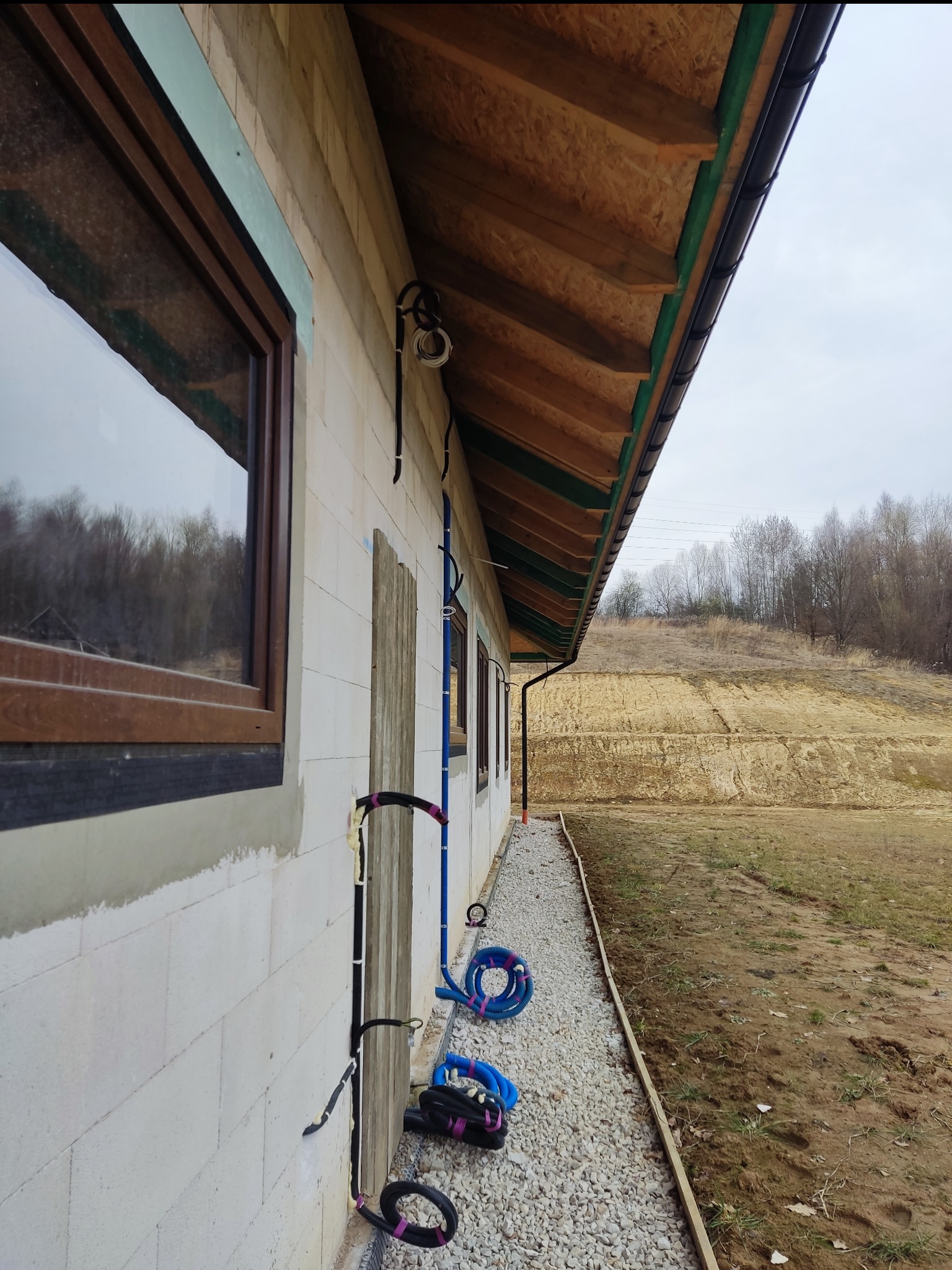 Instalacja elektryczna w budynku w trakcie budowy. Widoczne przewody prowadzone po elewacji z białego bloczka, okna z brązowymi ramami, rynna i podbitka dachowa.