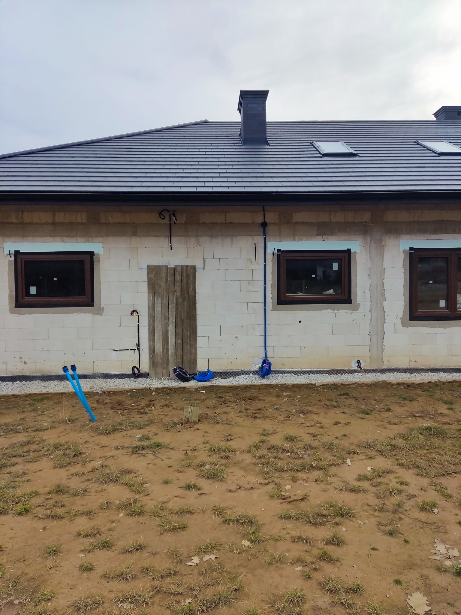 Surowy stan budynku z widocznymi przewodami i rurami instalacji na elewacji. Okna z brązowymi ramami, dachówka w kolorze grafitowym. Budynek w trakcie budowy.