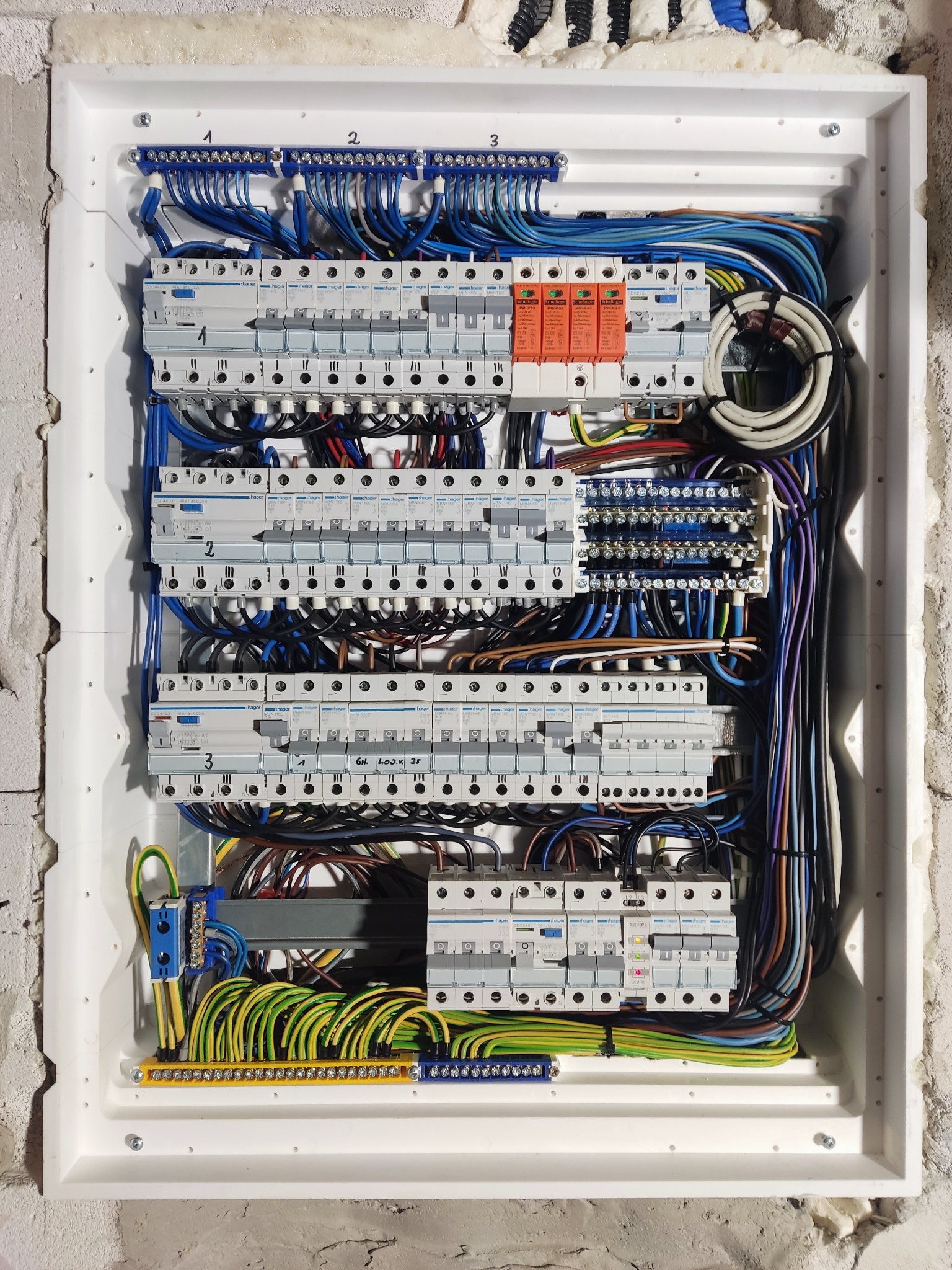 Rozdzielnica elektryczna w białej obudowie, widoczne bezpieczniki, przewody w różnych kolorach, zamontowana na ścianie z surowego betonu.