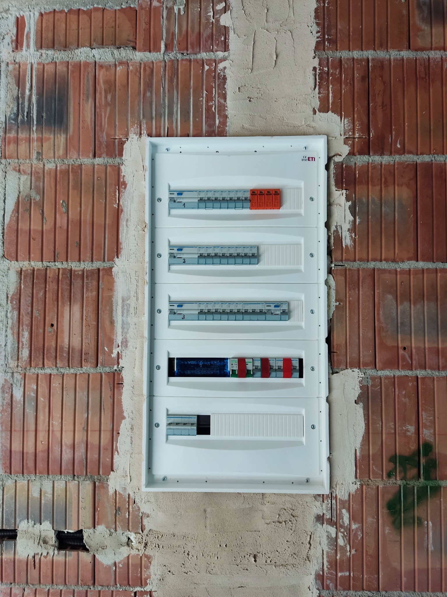 Rozdzielnica elektryczna ETI zamontowana w surowej ścianie z czerwonej cegły, widoczne zabezpieczenia i wyłączniki. Ściana w trakcie prac wykończeniowych.
