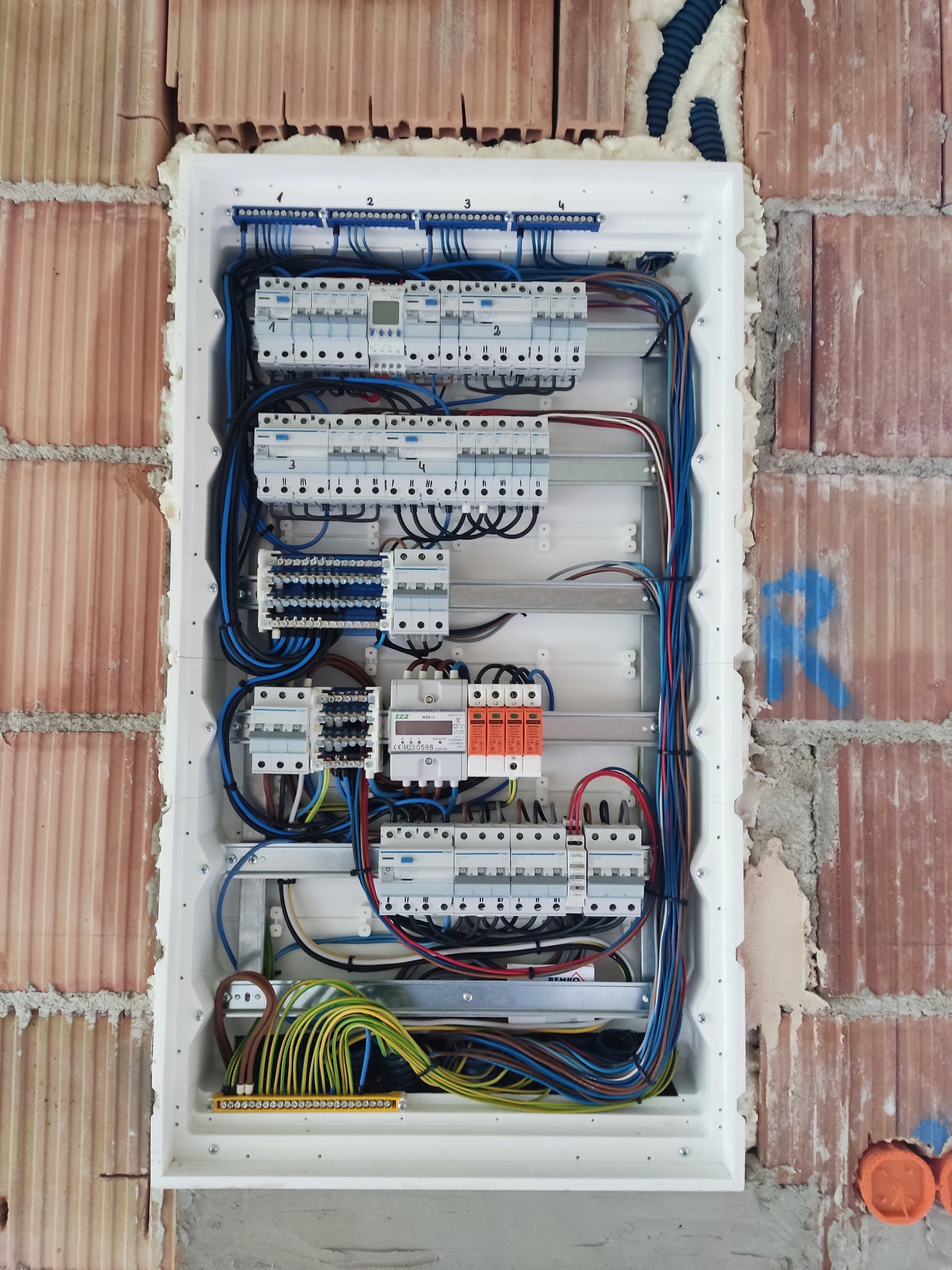 Rozdzielnia elektryczna w białej obudowie, zamontowana w surowej ścianie z cegły. Widoczne liczne przewody i zabezpieczenia. Instalacja w trakcie montażu.