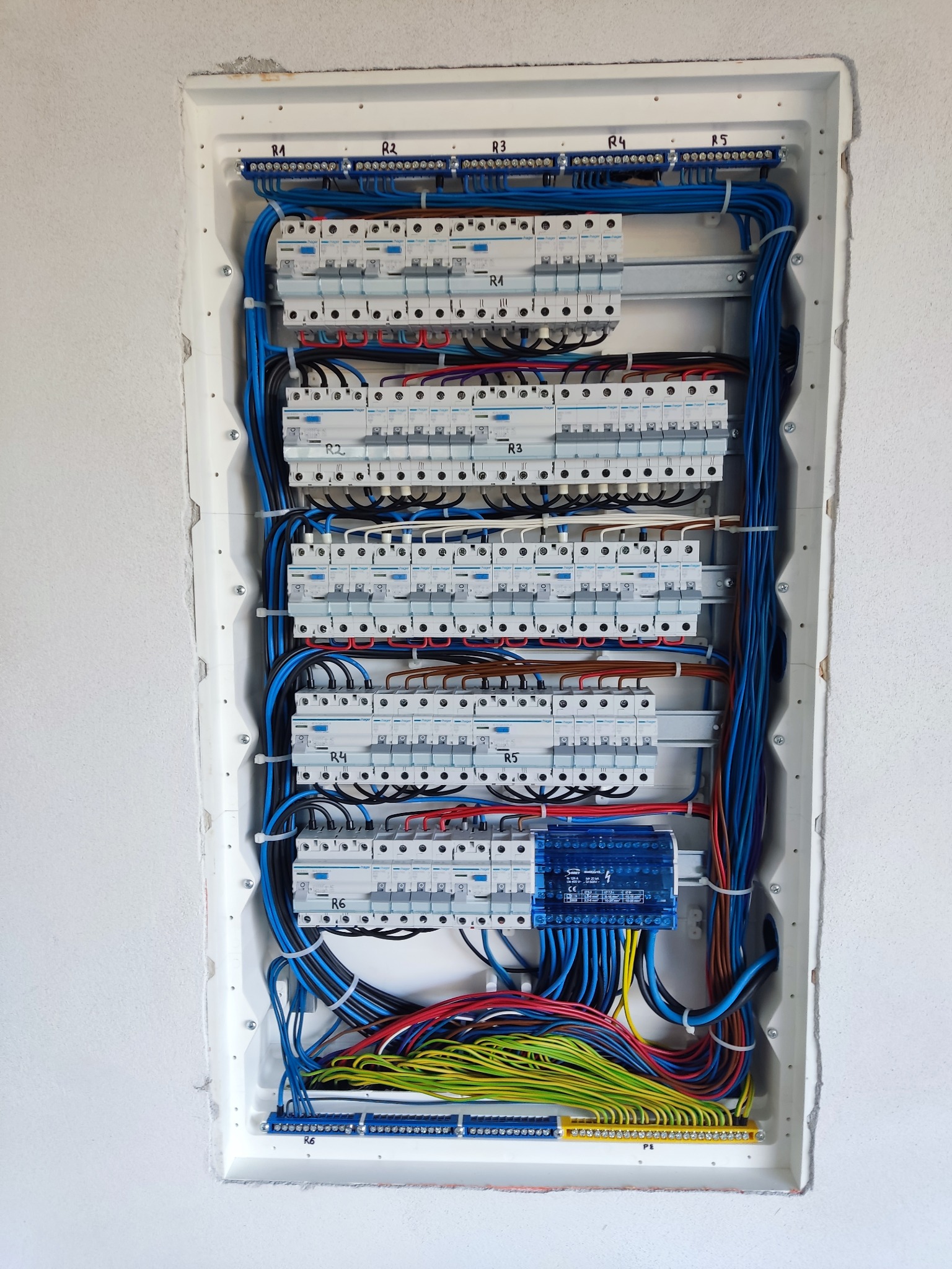 Rozdzielnica elektryczna Hager w białej obudowie, z widocznymi bezpiecznikami, przewodami o różnych kolorach i oznaczeniami R1-R6. Instalacja w surowym stanie.