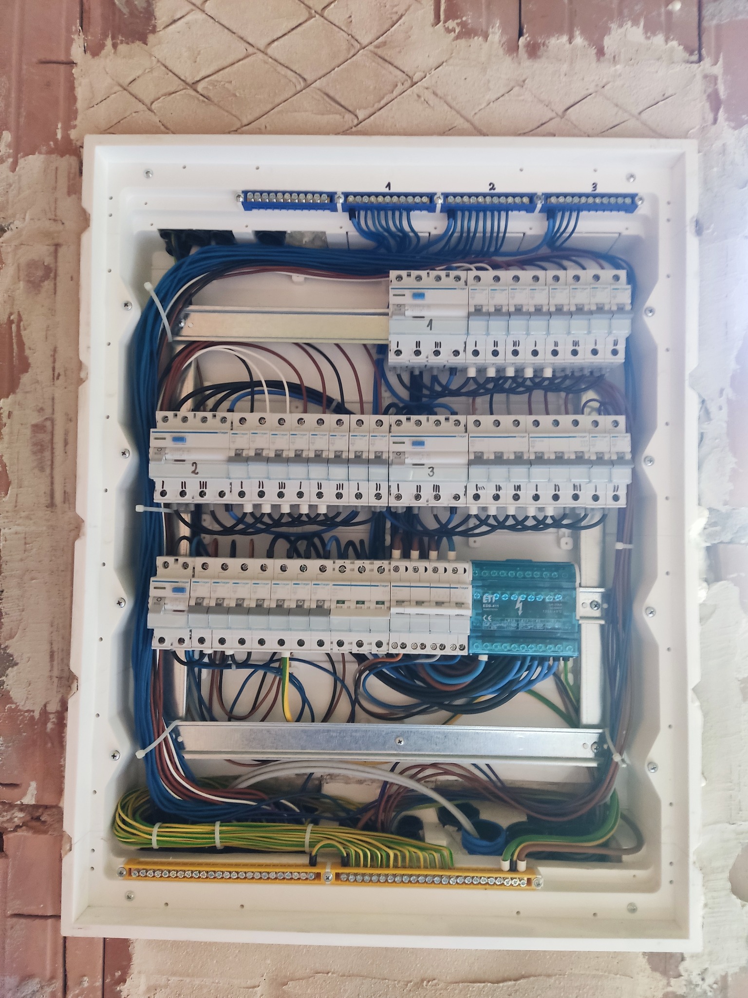 Rozdzielnica elektryczna w białej obudowie, z widocznymi bezpiecznikami, przewodami i listwami zaciskowymi, zamontowana na surowej ścianie z cegły.