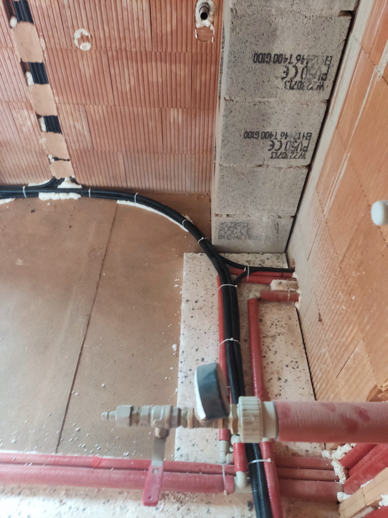 Układanie przewodów w ścianie z bloczków. Czarny peszel z kablami i czerwone rury instalacji wodnej/grzewczej przymocowane do ściany z cegły. Etap prac instalacyjnych.