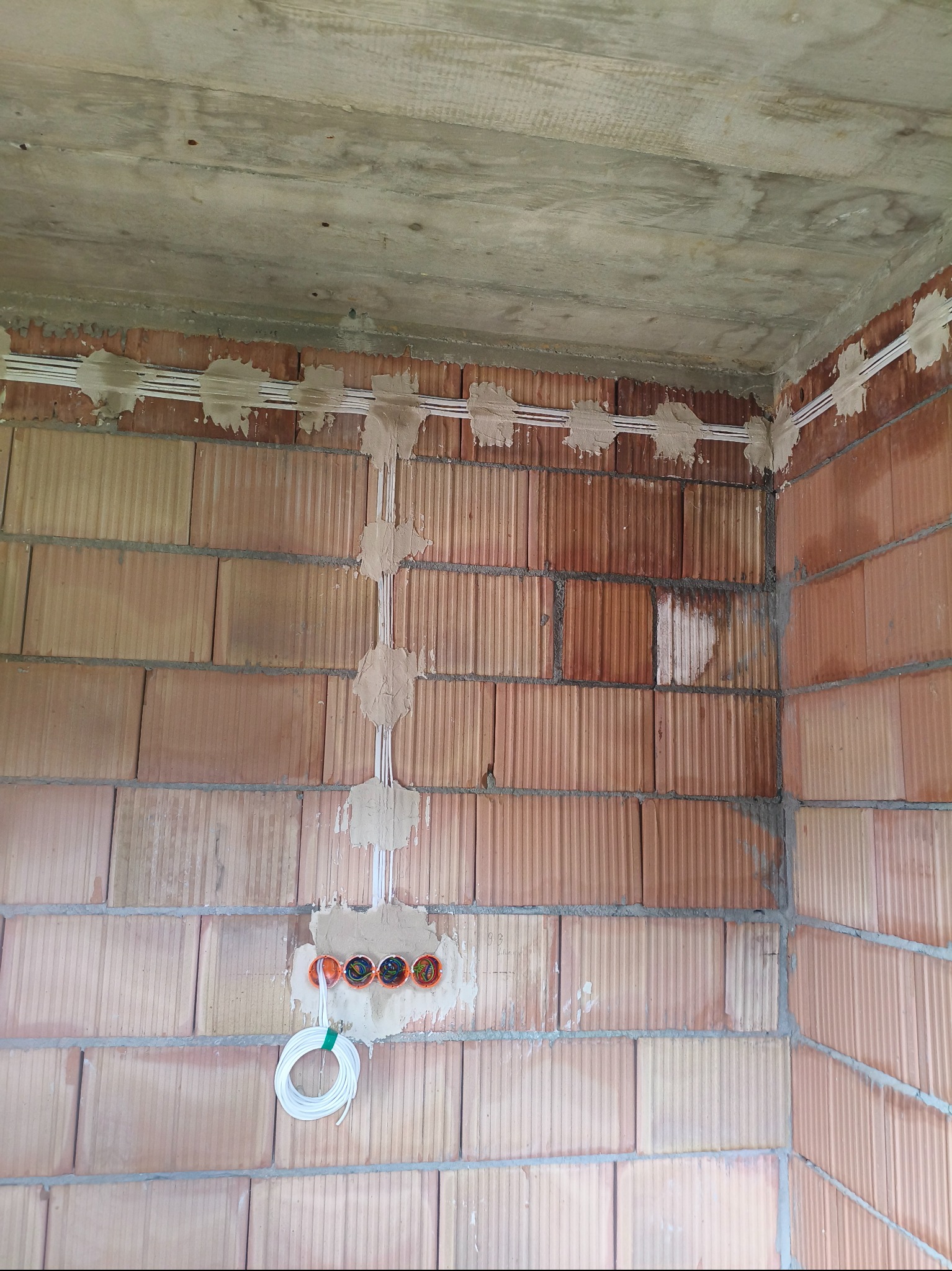 Wewnętrzne okablowanie w surowym wnętrzu z cegły. Widoczne kable przymocowane do ściany zaprawą, pomarańczowe puszki pod gniazdka i zwój kabla.