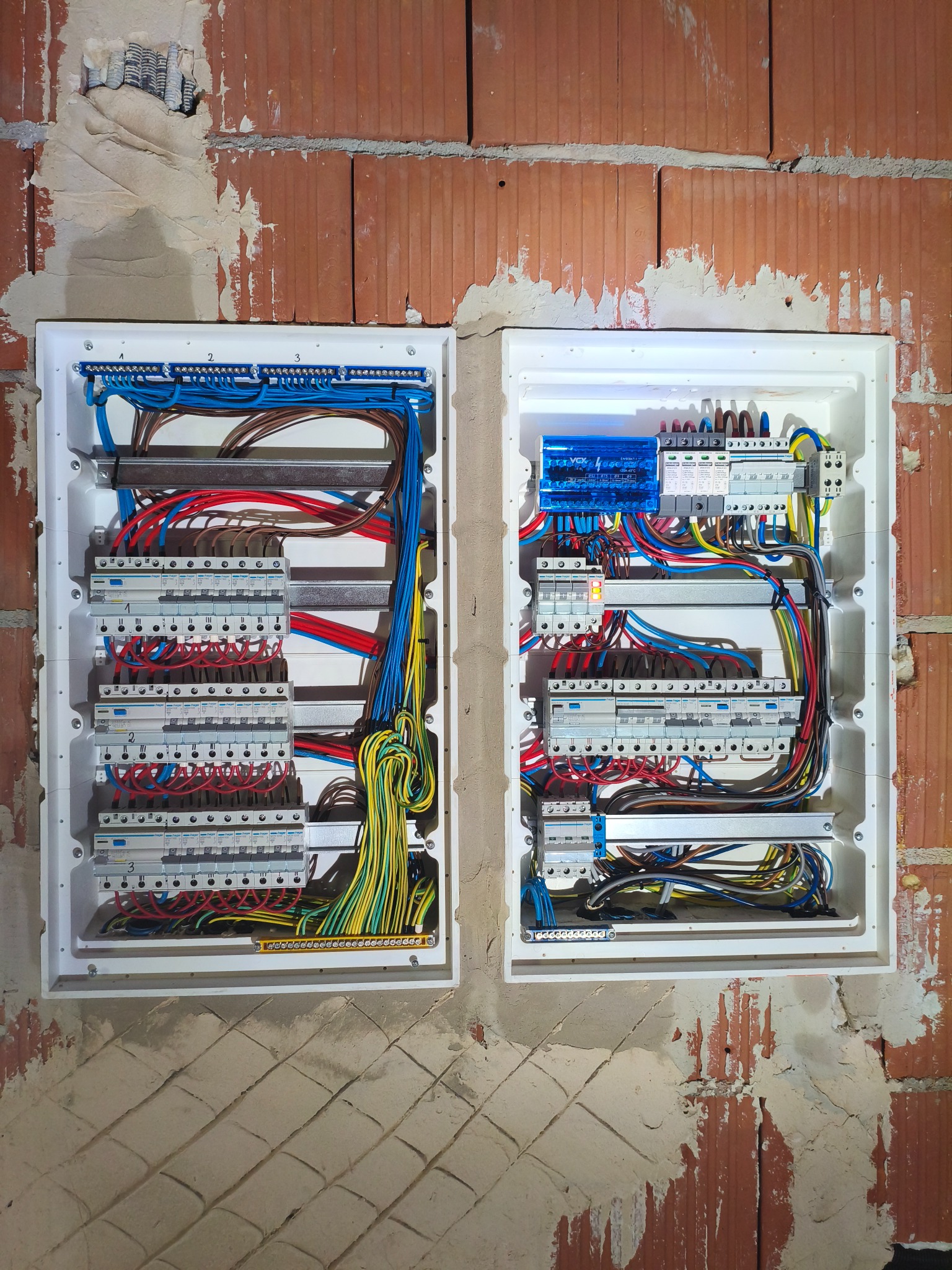 Dwie rozdzielnice elektryczne z okablowaniem zamontowane na surowej, ceglanej ścianie. Widoczne bezpieczniki, przewody w różnych kolorach i listwy zaciskowe.