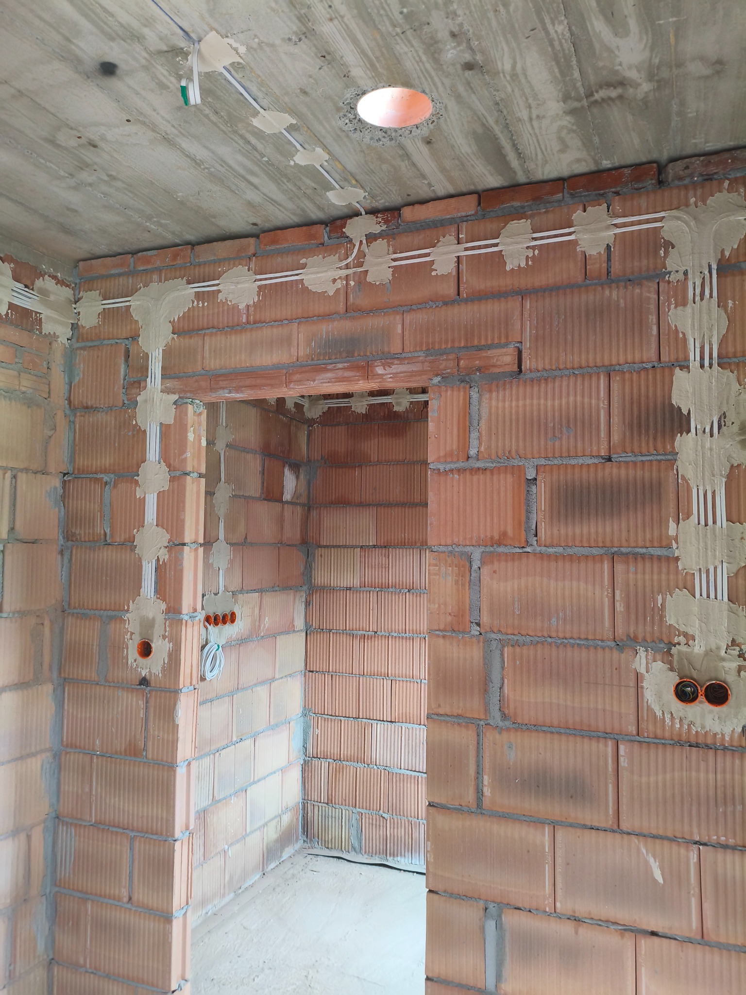 Wewnętrzne ściany z cegły z zamontowanymi przewodami elektrycznymi, zabezpieczonymi gipsem. Widoczne otwory na gniazdka i sufit z wywierconym otworem na lampę.