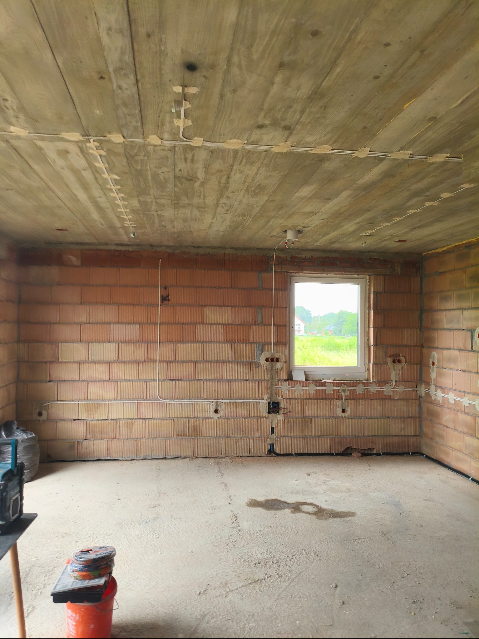 Wnętrze w stanie surowym z instalacją elektryczną: kable przymocowane do sufitu i ścian, widoczne puszki elektryczne, ceglany mur i surowy betonowy strop.