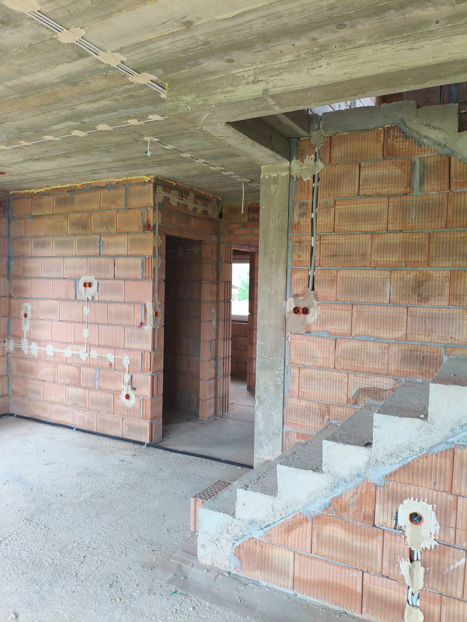 Wnętrze w trakcie instalacji elektrycznej: przewody w ścianach z cegły, puszki podtynkowe, kable na suficie, betonowe schody w surowym stanie. Widok na dwa pomieszczenia.