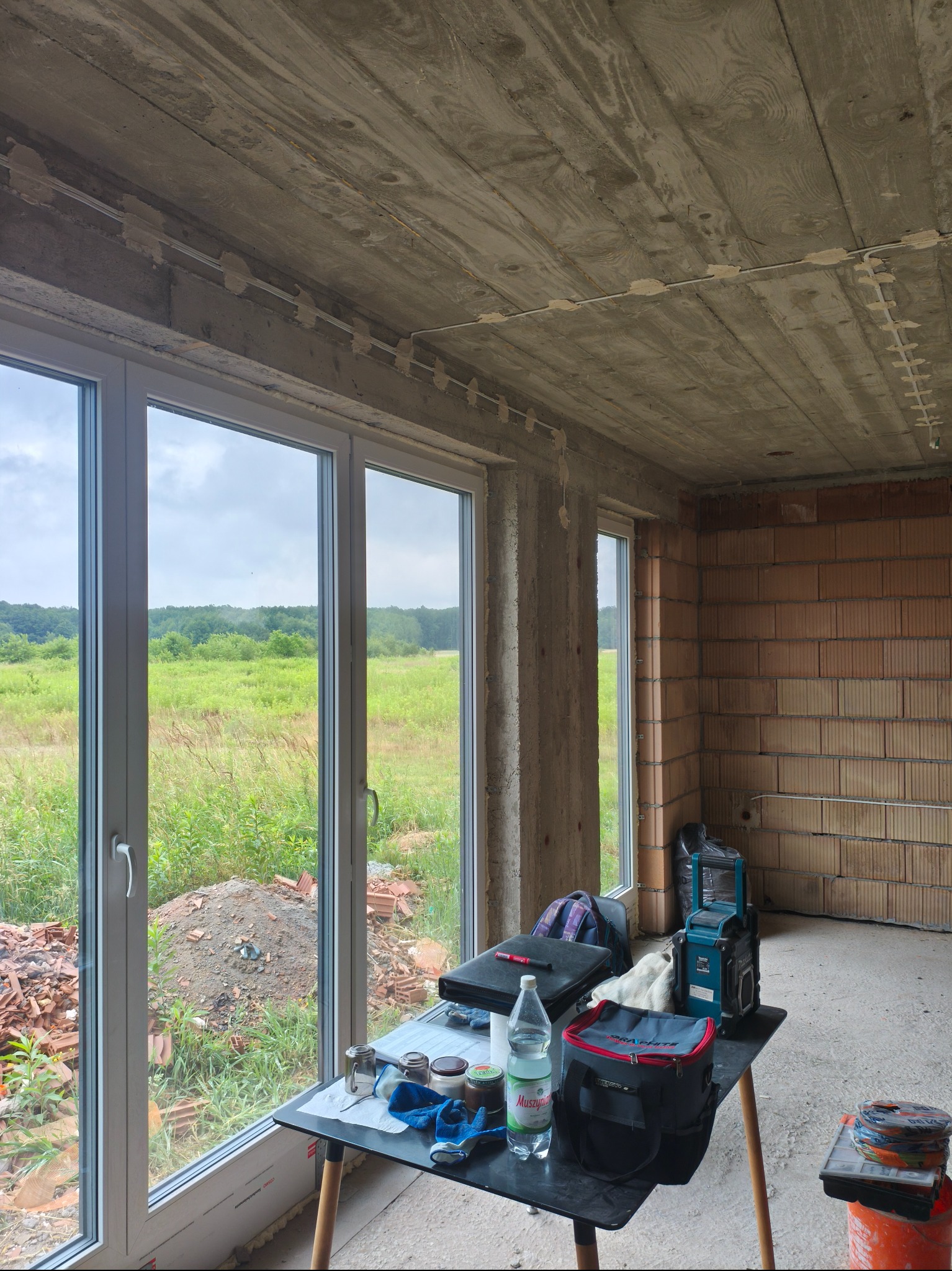 Wnętrze w stanie surowym z widocznymi przewodami elektrycznymi zamocowanymi na suficie. Na stole narzędzia i akcesoria elektryczne. Za oknem zielony krajobraz.