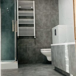 Usługi remontowe Gopus - Nowoczesna łazienka z szarymi płytkami, białym grzejnikiem drabinkowym na ścianie, kabiną prysznicową ze szklanymi drzwiami i podwieszaną toaletą.