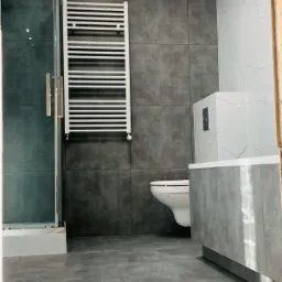 Nowoczesna łazienka z szarymi płytkami, białym grzejnikiem drabinkowym na ścianie, kabiną prysznicową ze szklanymi drzwiami i podwieszaną toaletą.