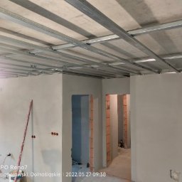 Usługi remontowe Gopus - Wnętrze w trakcie prac wykończeniowych: metalowa konstrukcja sufitu podwieszanego, ściany po wstępnym szpachlowaniu, widoczne otwory drzwiowe z surową cegłą, poziomica oparta o ścianę, instalacja...