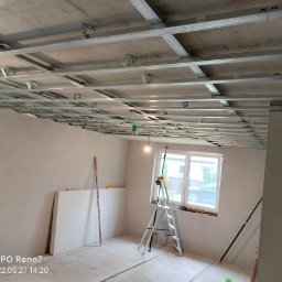 Usługi remontowe Gopus - Metalowa konstrukcja sufitowa w trakcie montażu, widoczne profile i zawiesia, w tle ściany przygotowane do dalszych prac wykończeniowych, drabina oparta o parapet okna.