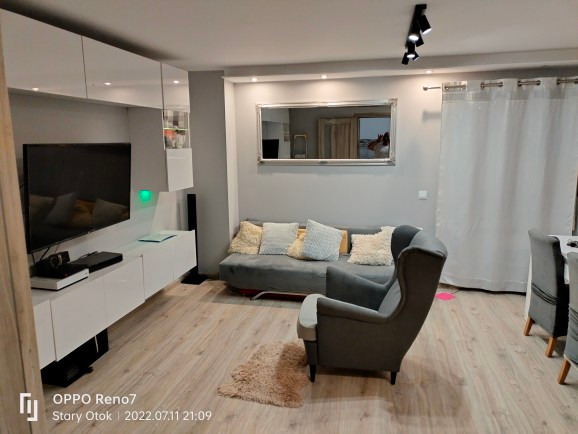 Jasne, nowocześnie urządzone wnętrze salonu z szarą sofą, fotelami i białymi meblami pod telewizorem. Na suficie widoczne czarne reflektory punktowe. Podłoga wyłożona panelami w odcieniach drewna.